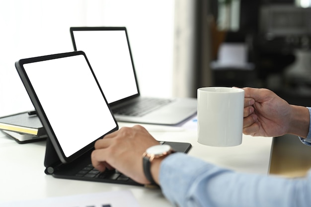 Toma recortada del empresario trabajando en la tableta de la computadora mientras sostiene la taza de café en la oficina.