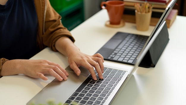 Toma recortada de la mano femenina escribiendo en el teclado de la computadora portátil en el escritorio de la oficina con otra computadora portátil, libros y artículos de papelería