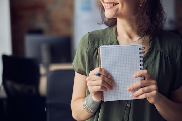 Foto toma recortada de mujer joven en ropa casual sosteniendo un cuaderno vacío y un bolígrafo en sus manos y sonriendo