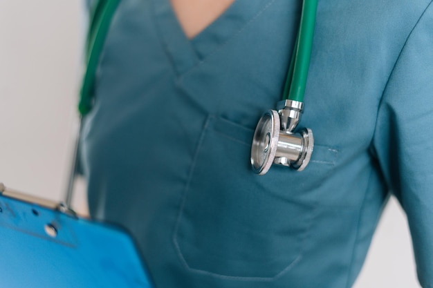 Toma recortada de primer plano de una doctora irreconocible con uniforme médico verde usando estetoscopio sosteniendo portapapeles, de pie sobre fondo blanco aislado en estudio, enfoque selectivo.