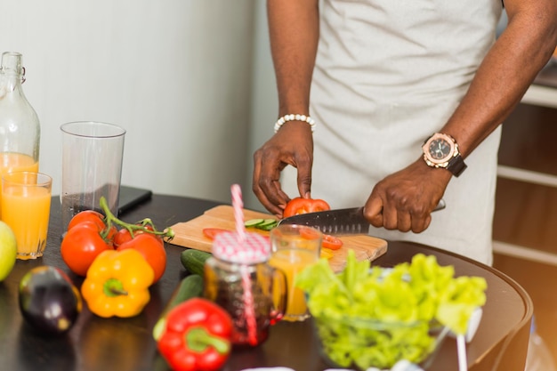 Toma recortada de primer plano de hombre africano preparando ensalada de verduras, sosteniendo un cuchillo, cortando tomates en una tabla de cortar en la cocina de casa. Concepto de comida vegetariana y saludable.