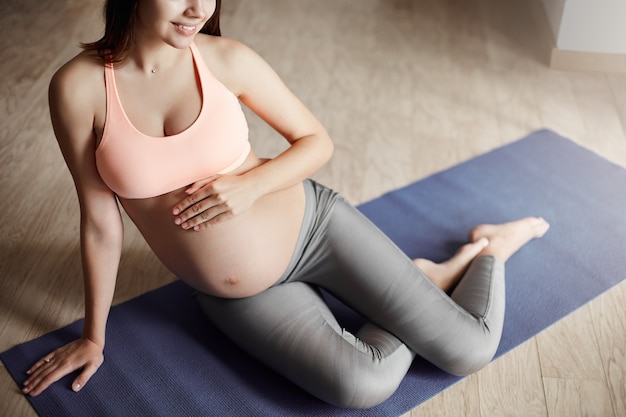 Toma recortada de tierna y cariñosa mujer embarazada en sujetador deportivo y leggings, acostada sobre una almohadilla antivuelco en la sala de estar, sonriendo ampliamente y tocando el vientre, sintiendo al bebé moviéndose hacia adentro, soñando con el futuro.