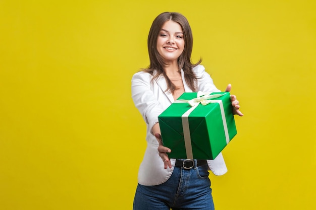 Toma este regalo. Retrato de una generosa morena con chaqueta y jeans sosteniendo un regalo y sonriendo a la cámara, feliz de compartir el regalo, la donación. tiro de estudio interior aislado sobre fondo amarillo