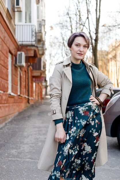 Toma de retrato de una atractiva mujer elegante atajo, niña camina en la ciudad al aire libre. Imagen elegante, moderna y femenina, estilo. Chica con capa o abrigo beige y vestido verde