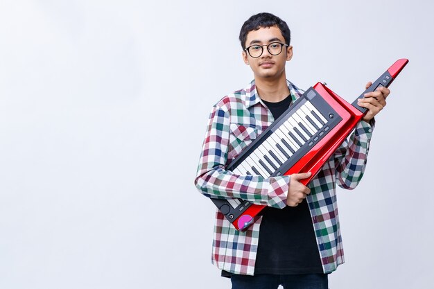 Toma de retrato de guapo joven músico adolescente tocando el keytar mientras mira a la cámara en el estudio. Joven músico junior de pie y sosteniendo keytar aislado con fondo blanco.