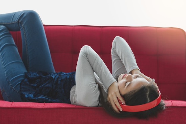 Toma de retrato de joven adolescente tailandés-turco acostado en el sofá rojo y disfrutando de escuchar música con un auricular rojo con las manos tocan el gesto de los auriculares. Concepto de afición y relajación.