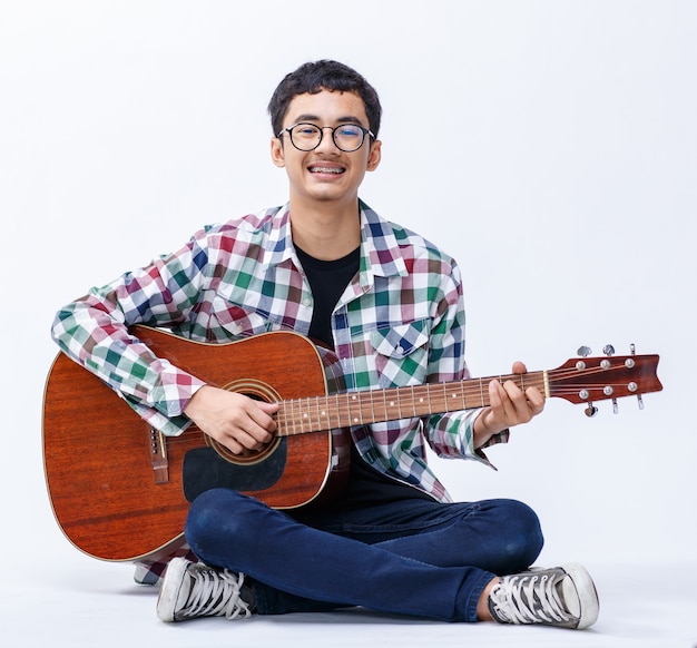 Toma de retrato de un lindo adolescente varón sonriente sosteniendo la guitarra acústica. Guitarrista junior profesional sentado y tocando un instrumento mientras mira a la cámara aislado fondo blanco.