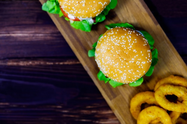 Toma de vista superior de cerca de dos deliciosas hamburguesas deliciosas sabrosas pan con tomate de res y lechuga sirven con aros de cebolla fritos en placa de madera decorada en tela de saco con perejil en la mesa de madera vieja oscura.