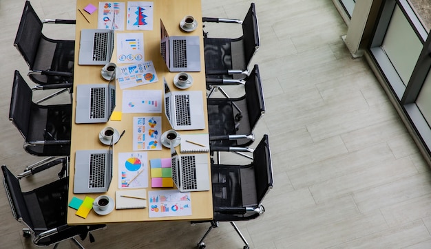 Toma de vista superior de la sala de reuniones de conferencias en la oficina de la empresa llena de computadoras portátiles en el escritorio de trabajo con sillas negras y monitor de pantalla grande y tablero de vidrio con documentos en papel de datos post-it.