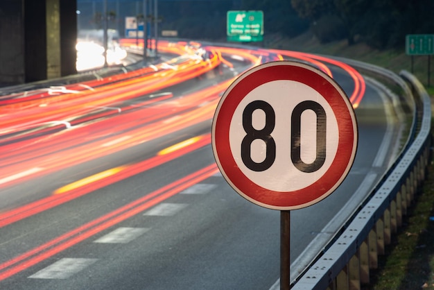 Tomada de exposición larga de una señal de tráfico que muestra el límite de velocidad de 80 km/h en una autopista llena de coches en movimiento borroso durante la noche