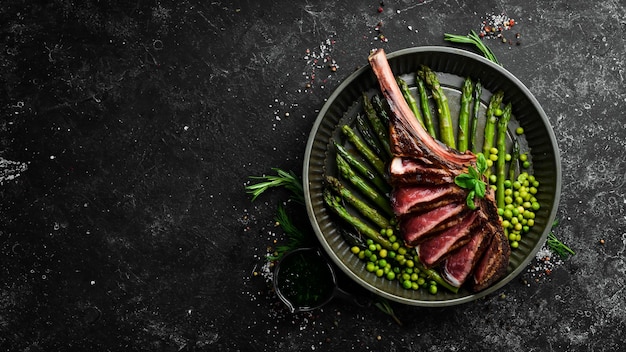 Foto tomahawk-steak am knochen gegrilltes steak mit spargel und grünen erbsen auf schwarzem hintergrund draufsicht freier kopierbereich