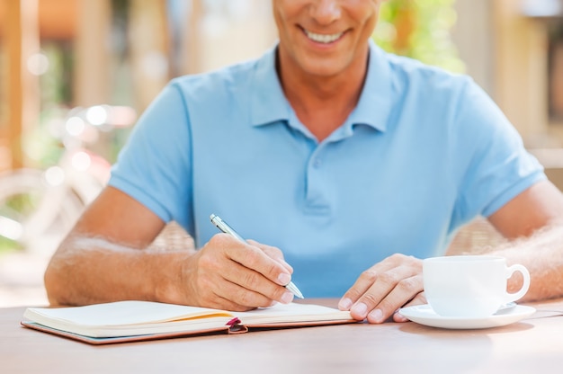 Tomando algunas notas importantes. Primer plano de hombre maduro seguro de escribir algo en su bloc de notas y sonriendo mientras está sentado en la mesa al aire libre con casa en el fondo