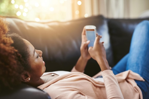 Tomando las cosas con calma Toma de ángulo alto de una mujer joven atractiva usando su teléfono celular mientras se relaja en el sofá de su casa