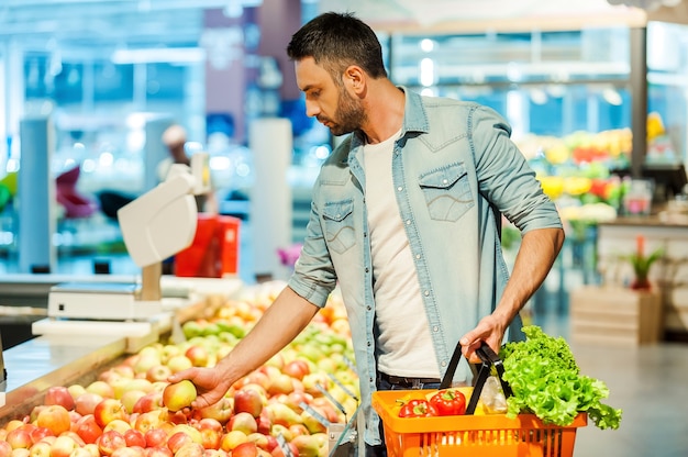 Tomando decisiones nutricionales Hombre joven seguro que sostiene la manzana y la bolsa de compras mientras está de pie en una tienda de alimentos