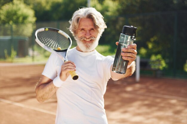 Tomando un descanso y bebiendo agua Senior hombre moderno y elegante con raqueta al aire libre en la cancha de tenis durante el día