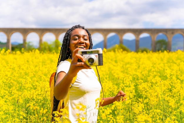Tomando fotos sonriendo con una cámara vintage una chica étnica negra con trenzas un viajero en un campo de flores amarillas