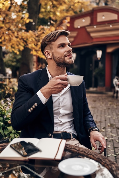 Tomando un momento para disfrutar del día. Apuesto joven sonriente en ropa casual elegante sosteniendo una taza de café y mirando a otro lado con una sonrisa mientras está sentado en el restaurante al aire libre