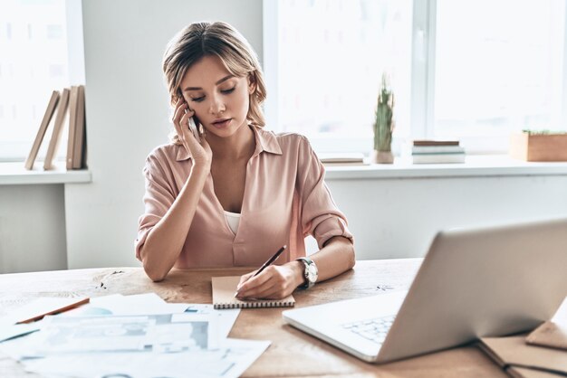 Tomando notas. Hermosa mujer joven en ropa casual elegante escribiendo algo y hablando por teléfono mientras está sentado en la oficina moderna