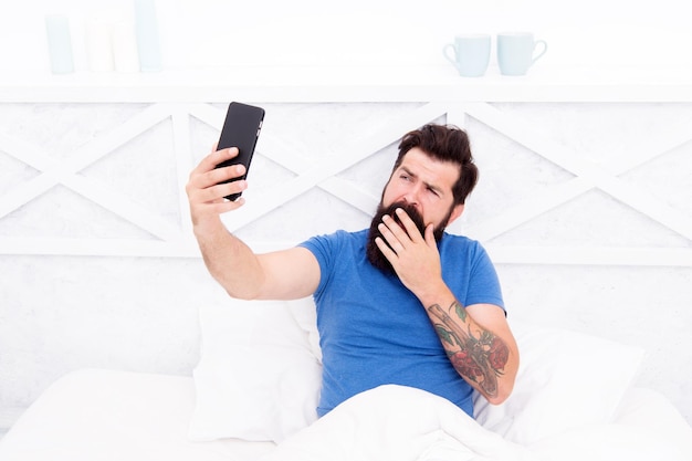 Tomando selfie Comunicación celular Chico moderno hablando por teléfono inteligente en la cama Tecnología moderna para la vida cotidiana Comunicación moderna Enviar foto Hombre guapo con teléfono móvil moderno desde casa