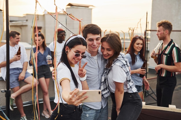 Tomando selfie Grupo de jóvenes con ropa informal tienen una fiesta en la azotea juntos durante el día