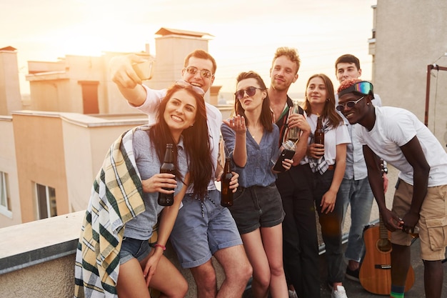 Tomando selfie Hermoso sol Grupo de jóvenes con ropa informal tienen una fiesta en la azotea juntos durante el día