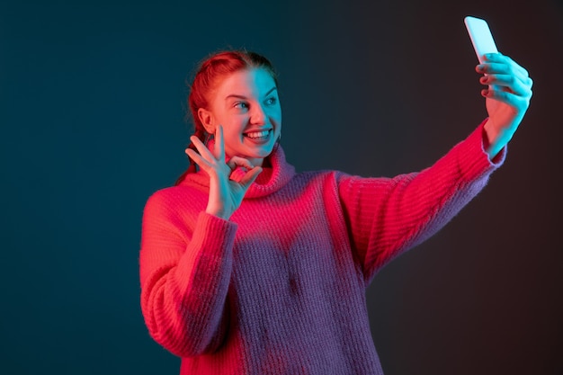 Tomando selfie, vlog, videollamada. Retrato de mujer caucásica aislado sobre fondo de estudio degradado en luz de neón. Modelo de mujer hermosa con el pelo rojo. Concepto de emociones humanas, expresión facial, anuncio.