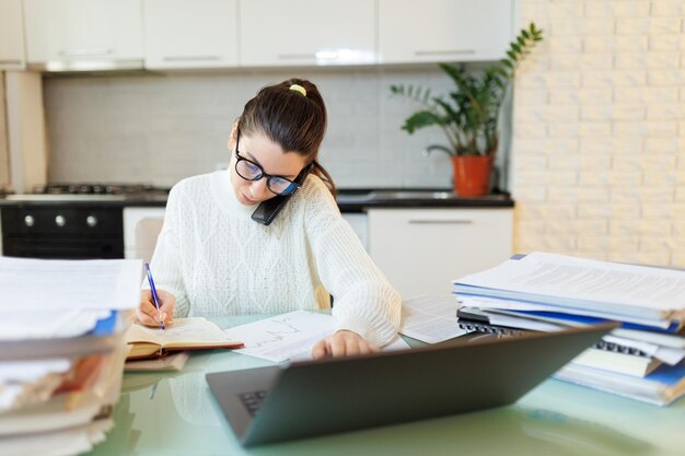 Tomando uma pausa uma mulher trabalha em casa em seu laptop falando ao telefone com documentos espalhados