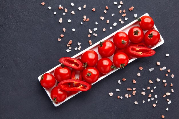 Tomate cereja lavado fresco e fatias de páprica em um prato oblongo branco com sal rosa em uma mesa de concreto preto