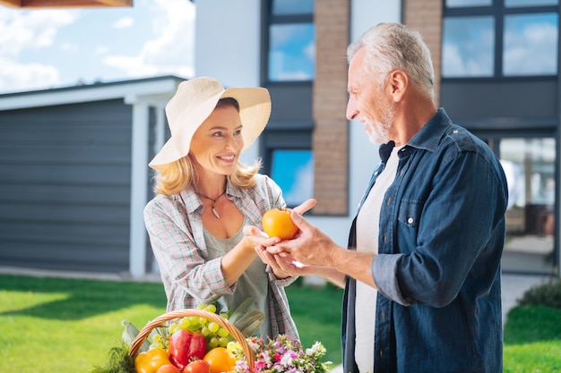 Tomate fresco. Cuidado esposa rubia vistiendo elegante sombrero beige dando tomate fresco agradable a su marido barbudo