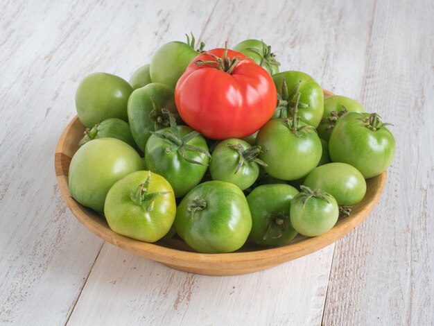 Un tomate rojo entre varios tomates verdes en una placa de madera.