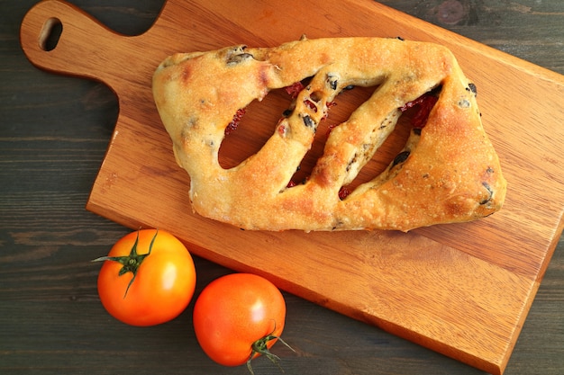 Foto tomate seco ao sol e pão francês de azeitona fougasse na bandeja de madeira com dois tomates frescos