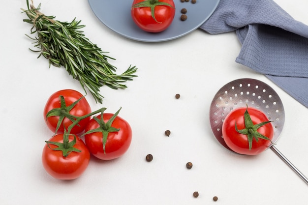 Tomaten auf grünem Zweig. Tomate und Piment auf grauem Teller. Tomate mit grünem Schwanz am Abschäumer. Graue Serviette. Weißer Hintergrund. Flach liegen