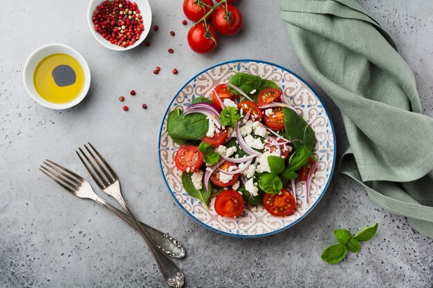 Tomaten, Spinatblätter, rote Zwiebeln und Feta-Käse-Salat auf hellem Keramikteller auf grauem Hintergrund