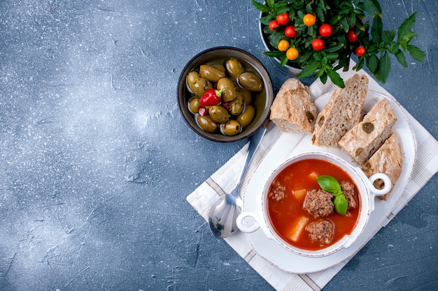 Tomatensuppe in einem weißen Teller mit Fleischbällchen, auf einem Stein Hintergrund mit Brot und Oliven