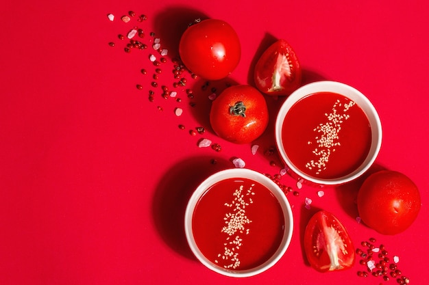 Tomatensuppe mit Sesamsamen in Schalen auf rotem Grund. Reifes Gemüse, rosa Himalayasalz, rosa Pfefferkörner. Ein modernes hartes Licht, dunkler Schatten, Draufsicht