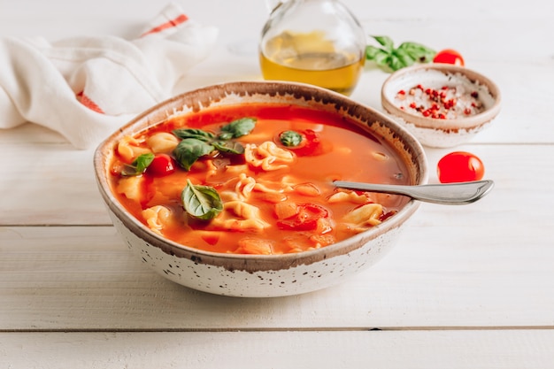 Tomatensuppe mit Tortellini in der weißen Schüssel auf weißem Holztisch.