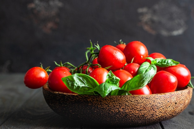 Tomates cherry y albahaca en un plato de madera sobre una superficie oscura.