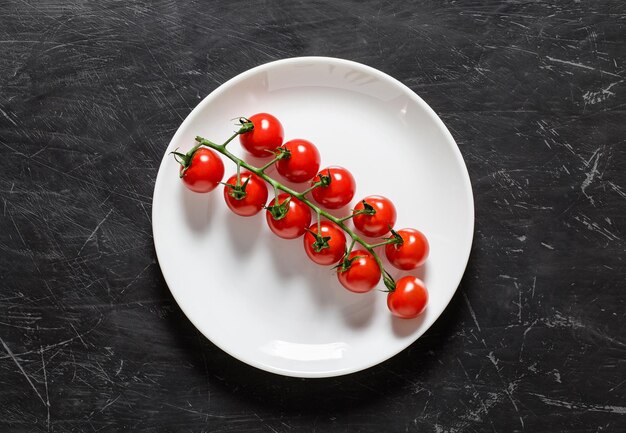 Tomates cherry, rama fresca en plato blanco, fondo oscuro de vista superior.