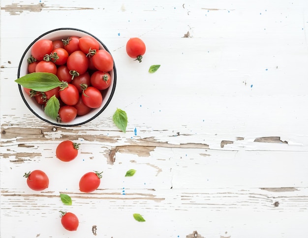 Tomates cherry en tazón de metal y hojas de albahaca fresca en la vista superior de fondo de madera blanca rústica