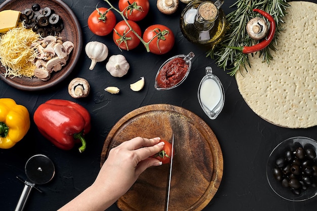 Tomates cortados a mano femenina en una tabla de madera en la mesa de la cocina alrededor de los ingredientes para pizza vegetal