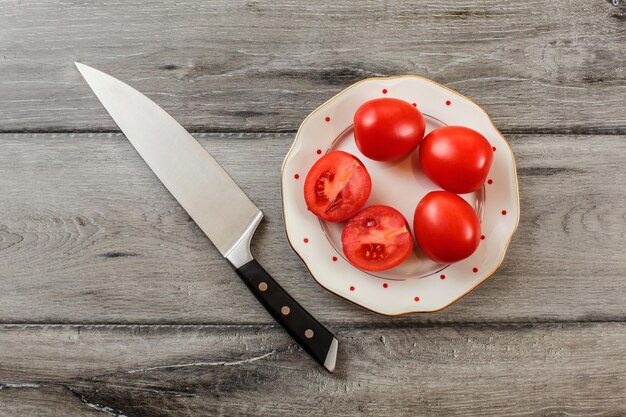 Tomates, uno de ellos cortado por la mitad en un plato de porcelana blanca con pequeños puntos rojos, cuchillo de chef tirado cerca. Vista de mesa.