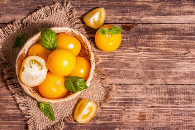 Tomates maduros amarelos com manjericão fresco. Novos vegetais de colheita em uma cesta de vime. Mesa de madeira vintage, vista superior