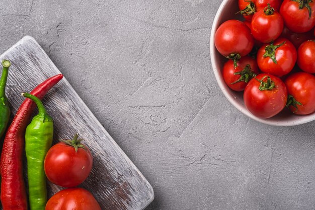 Tomates maduros frescos en un tazón cerca de la vieja tabla de cortar de madera con pimientos picantes, espacio de copia de hormigón de piedra