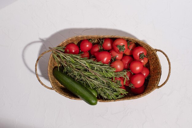 Tomates, pepinos y unas ramitas de romero en una canasta de mimbre sobre una superficie blanca