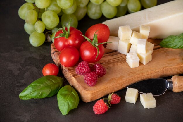 Tomates, queso, albahaca, frambuesas y uvas en una tabla de olivo sobre un fondo oscuro