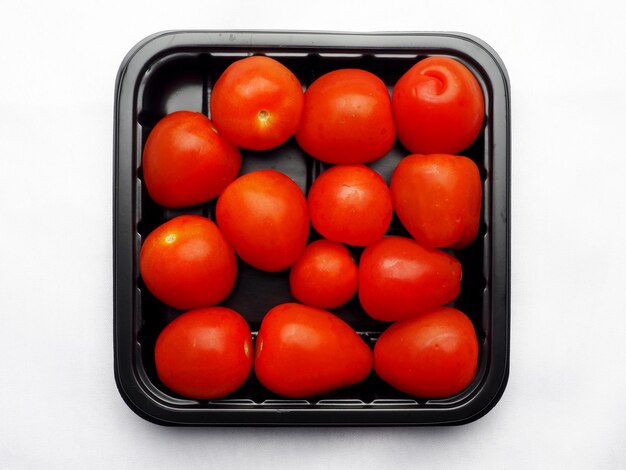 Tomates vermelhos frescos de tamanho pequeno em recipiente de plástico preto sobre fundo branco, ingrediente alimentar colorido para cozinhar