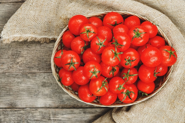 Tomates vermelhos frescos em uma cesta de vime em uma tabela de madeira velha. Tomates de cereja maduros e suculentos com gotas da umidade, tabela de madeira cinzenta, em torno de um pano da serapilheira. Em estilo rústico.