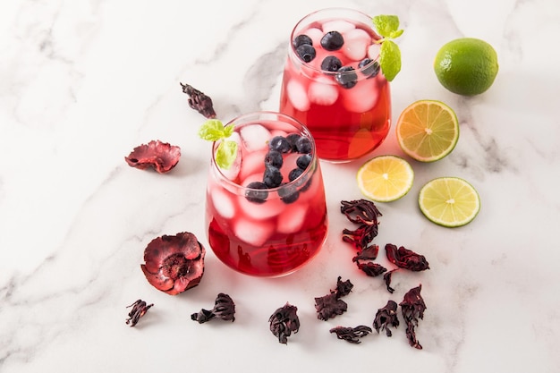 Tonischer Vitamin-Eistee-Rosenblüten-Hibiskus mit Heidelbeeren, Limettenstücken und Eiswürfeln in zwei Gläsern auf einem Marmortisch