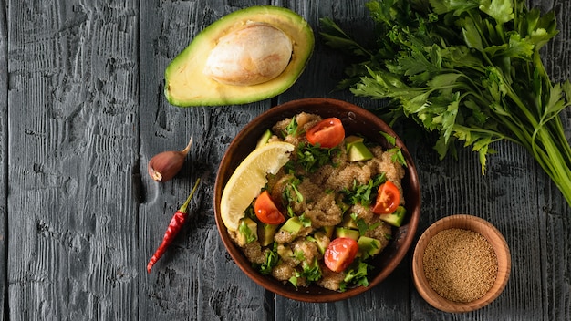 Tonschale mit Amaranthsalat mit Avocado, Zitrone, Tomaten und Petersilie und Schüssel mit Olivenöl auf einem dunkelschwarzen Tisch. Der Blick von oben.