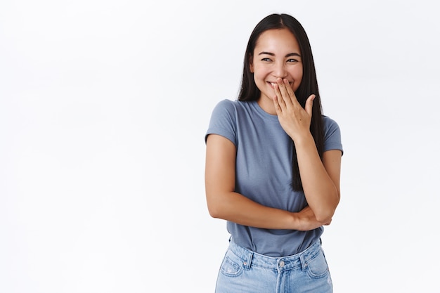 Tonto y horrible estudiante universitaria asiática haciendo una broma y riendo en un rincón mientras ve a alguien ser engañado, mirar a la izquierda, taparse la boca sonriente con la mano y reír secretamente, pared blanca